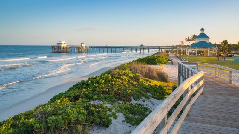 A wooden boardwalk leads toward a sandy beach with a long pier extending into the ocean under a clear sky.
