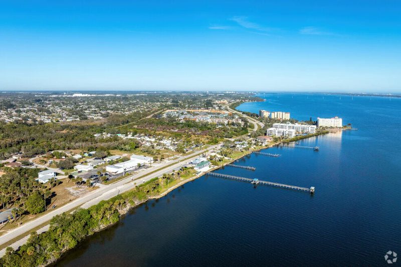Aerial view of a coastal road and waterfront homes with long wooden piers stretching into blue water.