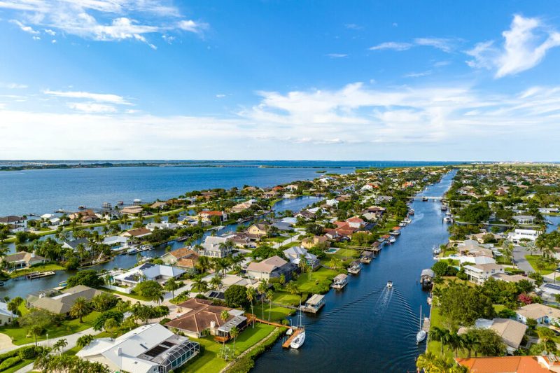 Aerial view of waterfront houses along a narrow canal leading out to a large bay under a bright blue sky.